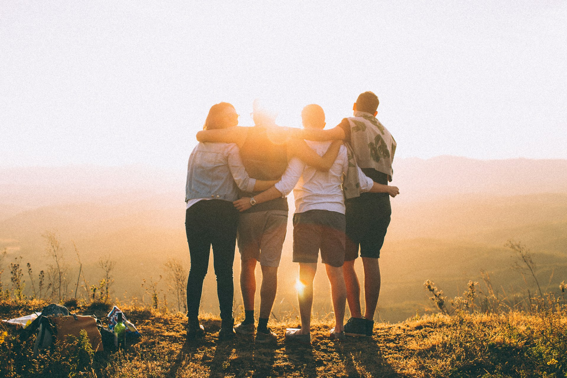 four people, shown from behind, with their arms around each other, facing a sunset or sunrise